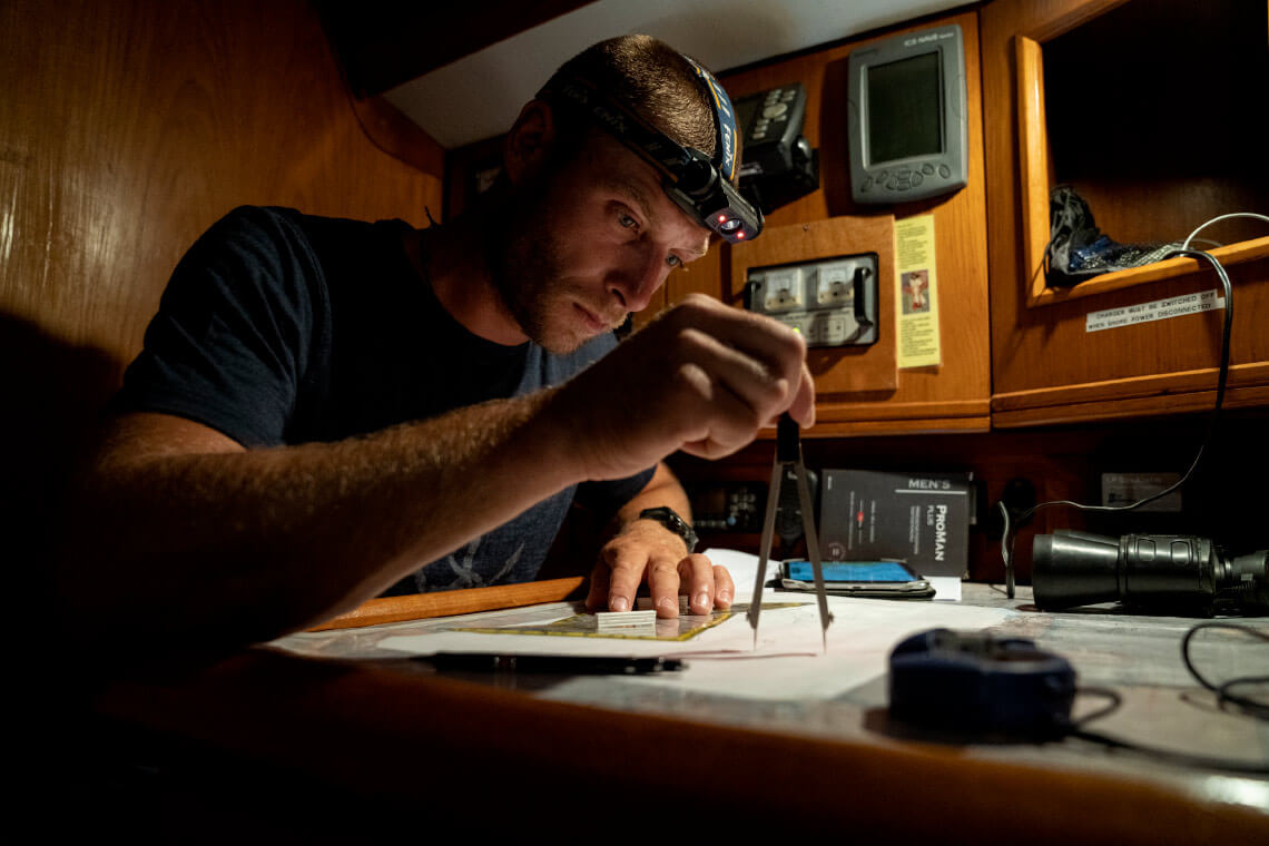 Natural Pharmaceuticals ProMan Plus Vyras su galvos žibintuvėliu kruopščiai braižo navigacijos planą jachtos kabinoje prie stalo. Ant stalo yra navigacijos priemonės, įskaitant kompasą, žemėlapius ir liniuotę. Fone matosi jachtos langai, pro kuriuos matosi tamsi nakties jūra.