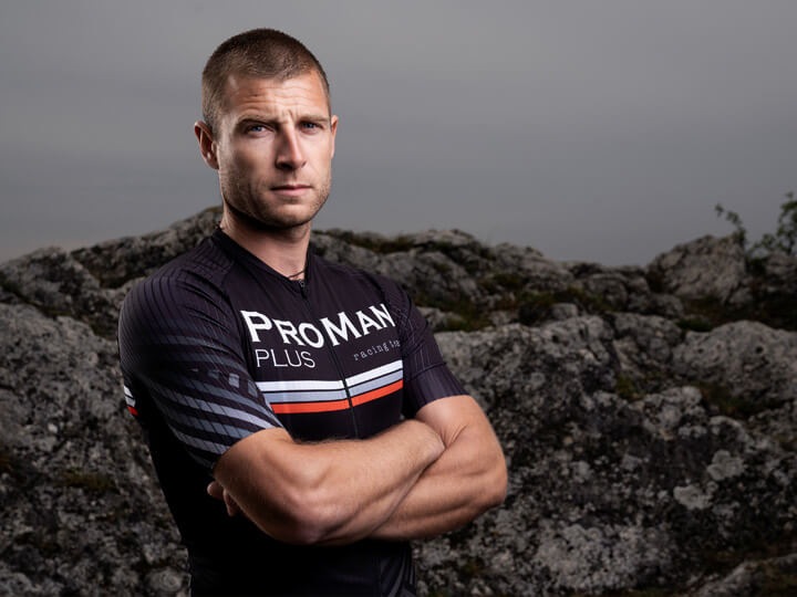 Natural Pharmaceuticals ProMan Plus Trumpaplaukis vyras, apsirengęs sportine apranga ir dėvintis „ProMan Plus“ dviračių aprangą, stovi susikaupęs prieš akmeninį foną.