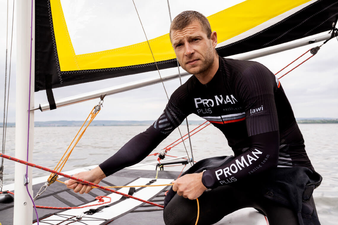 Natural Pharmaceuticals ProMan Plus Vyras sportine apranga su „ProMan Plus“ logotipu, vairuojantis burlaivį. Jis sėdi ant denio, laikydamas burės virves, o fone matosi vanduo ir horizontas. Ant jo aprangos matosi užrašai „ProMan Plus“ ir „racing team“ (lenktynių komanda).