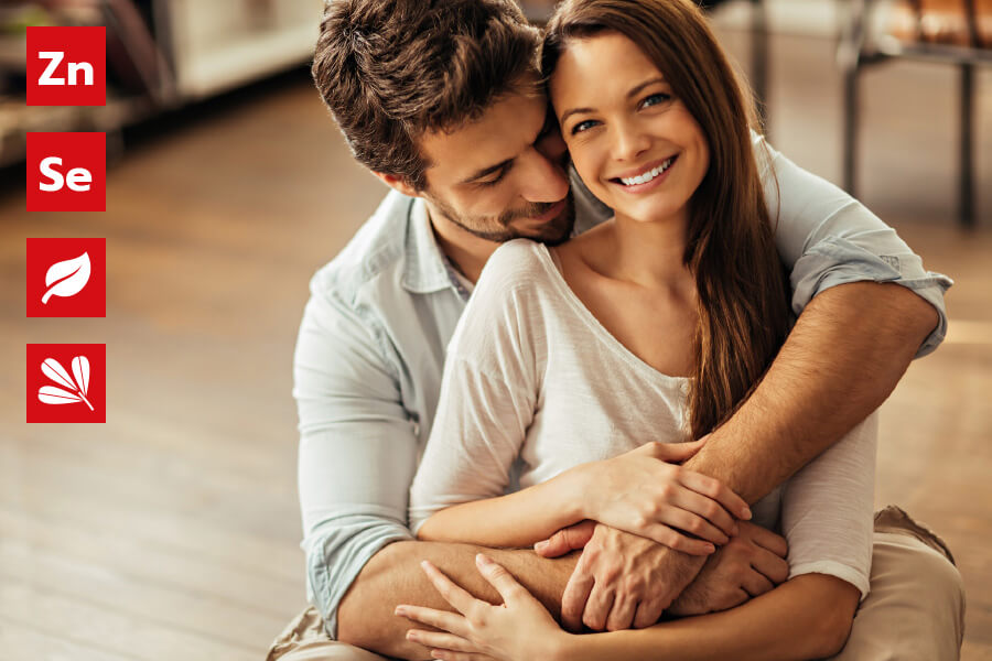 Natural Pharmaceuticals ProMan Plus Šypsosi pora, švelniai apsikabinusi. Kairėje – keturios raudonos piktogramos, simbolizuojančios sudedamąsias dalis: cinką (Zn), seleną (Se), natūralius augalų ekstraktus.